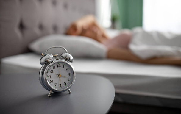 Hälytyskello yöpöydällä ja hämärä näkymä tuhatvuotisesta naisesta makaamassa sängyllä, haluton nousemaan ylös aamulla - Valokuva, kuva