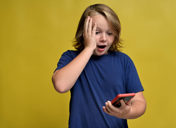 surprised cute teenager in blue t-shirt with long hair looks at the phone on a yellow background - Foto, Bild