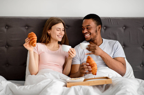 Multiraciaal stel samen ontbijten op bed, koffie drinken met croissants, huwelijksreis doorbrengen in hotel - Foto, afbeelding