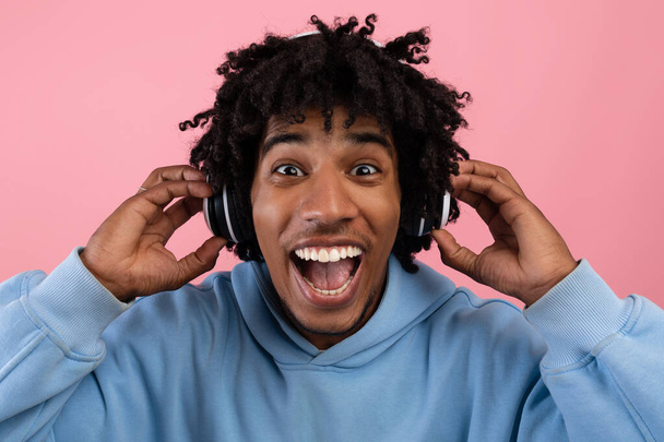 Adolescente negro lleno de alegría disfrutando de la música popular en los auriculares, la apertura de la boca en la emoción en el fondo rosa - Foto, Imagen