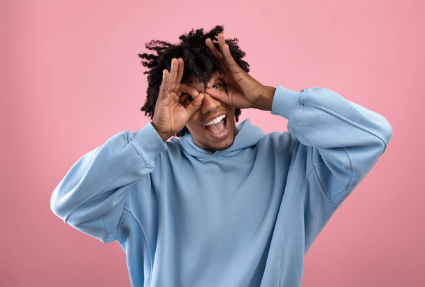 Silly black teen guy doing airplane glasses with his fingers, grimacing, making funny face on pink studio background - 写真・画像