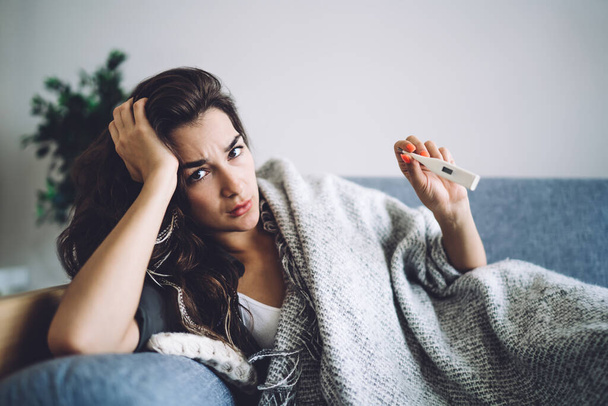 Tired caucasian female exhausted with virus disease lying on sofa checking temperature using modern termometr, sad woman stay at home feeling unwell catching covid-19 coronavirus care about health - Foto, imagen