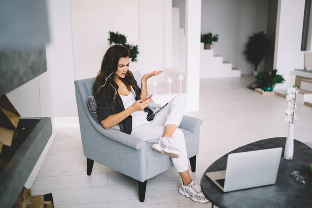 Confused caucasian female checking notification on mobile phone shocked with bill from banking app, 20s serious pensive woman checking messages on mobile phone spending free time in apartment - Photo, Image