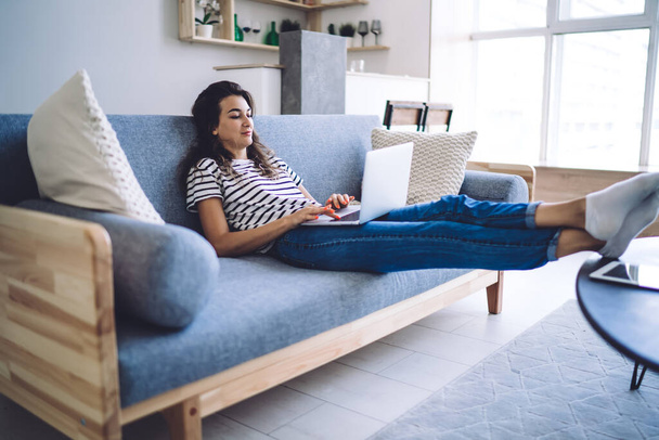 Young caucasian hipster girl relax at home interior watching movies on laptop computer lying on sofa, beautiful teen female in casual wear checking social media news spending free time at apartment - Photo, Image