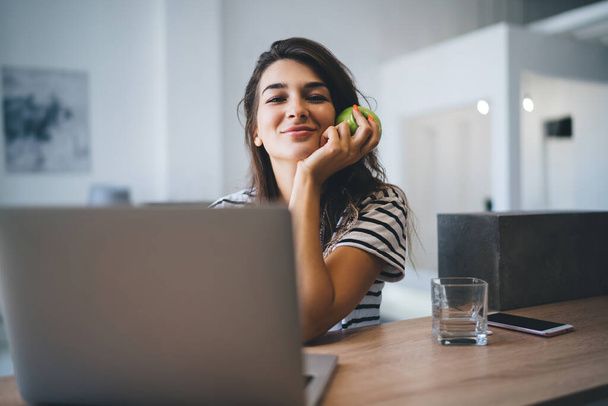 Half length portrait of cheerful caucasian female looking at camera holding apple waiting for food delivery making order online via laptop computer, successful woman freelancer enjoying remote job - Foto, afbeelding