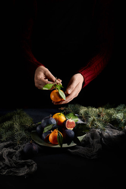 Dark Mood Christmas Composition with Fruit Plate and Christmas Tree Branch. Vintage Table with Purple Figs and Tangerine on Dark Green Plate. Man in Red Jersey holding tangerine - Foto, Imagen