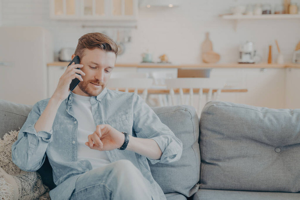 Mladý muž mluví po telefonu se svým přítelem, zatímco sedí na gauči - Fotografie, Obrázek