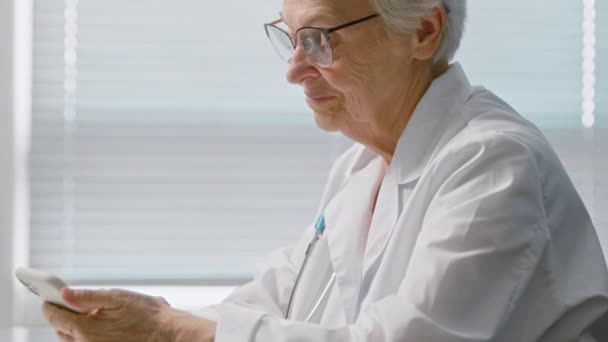 Mensaje de texto del médico en el celular sentado en la mesa en la clínica - Metraje, vídeo