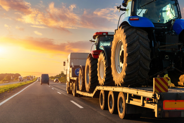 POV重工業トラックセミトレーラーフラットベッドプラットフォームは、日没の日の出の空で一般的な高速道路の道路に2つの大きな近代的な農業トラクターマシンを輸送します。農業設備輸送サービス - 写真・画像