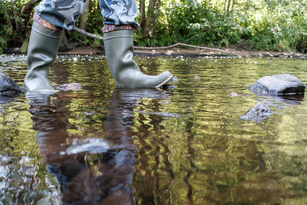 Los pies masculinos en botas de goma impermeables caminan a lo largo del río, contra el telón de fondo de la vegetación, en un día de verano. - Foto, imagen