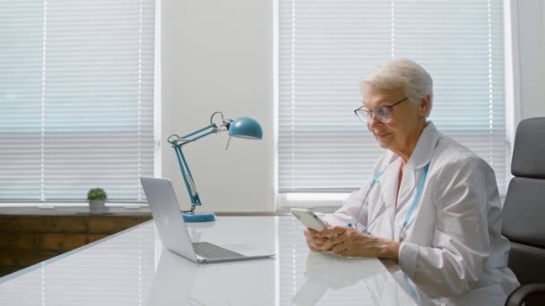 Hymyilevä lääkäri kirjoittaa viestin älypuhelimeen läppärin lähellä - Materiaali, video
