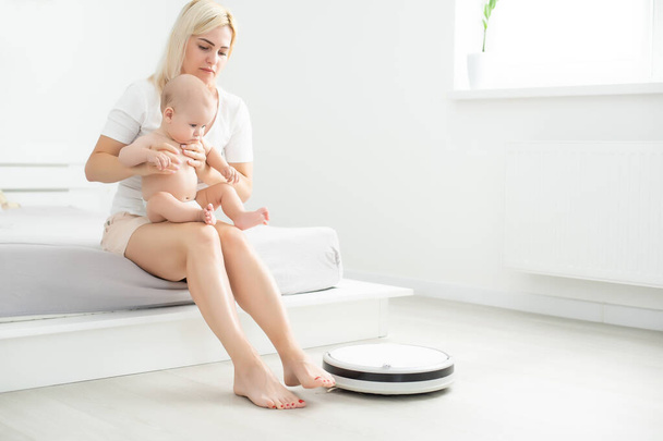 Young mother sitting on sofa with toddler son and watching robot vacuum cleaner doing housework. - Photo, Image