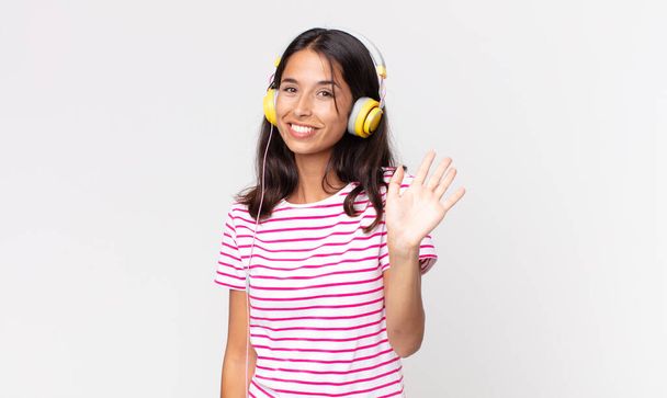 young hispanic woman smiling happily, waving hand, welcoming and greeting you listening music with headphones - Foto, immagini