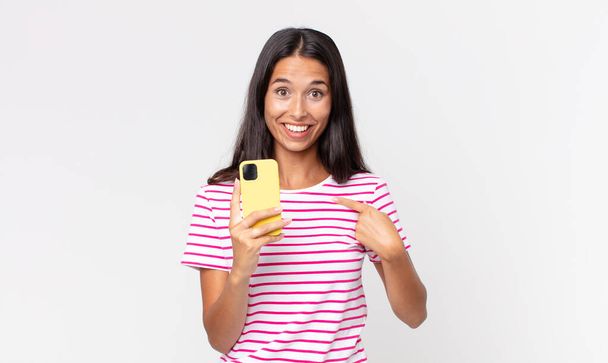 young hispanic woman feeling happy and pointing to self with an excited and holding a smartphone - Valokuva, kuva