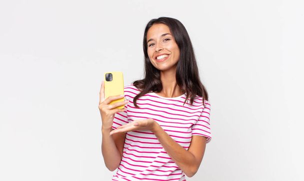 young hispanic woman smiling cheerfully, feeling happy and showing a concept and holding a smartphone - Zdjęcie, obraz