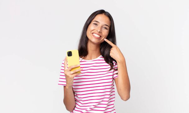 young hispanic woman smiling confidently pointing to own broad smile and holding a smartphone - Zdjęcie, obraz