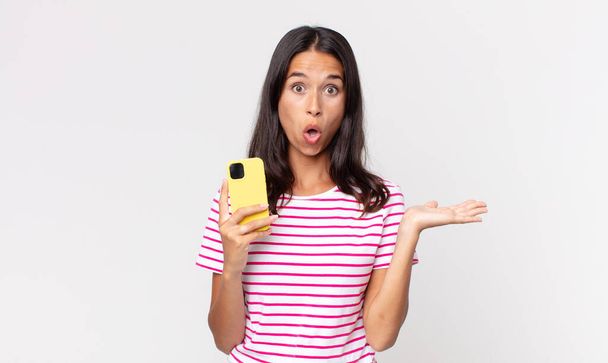 young hispanic woman looking surprised and shocked, with jaw dropped holding an object and holding a smartphone - Photo, image