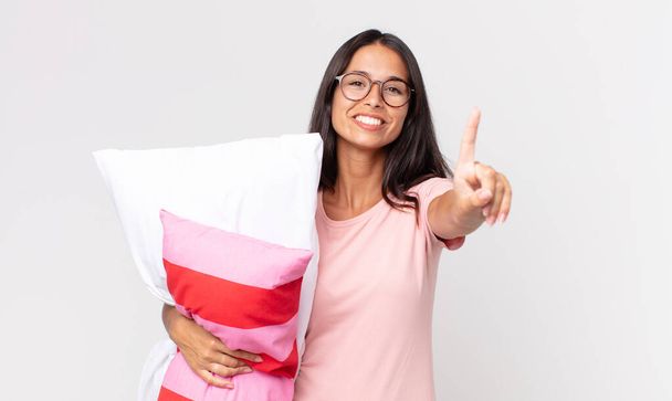 young hispanic woman smiling and looking friendly, showing number one wearing pajamas and holding a pillow - Foto, Imagem