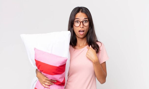 young hispanic woman looking shocked and surprised with mouth wide open, pointing to self wearing pajamas and holding a pillow - Foto, imagen