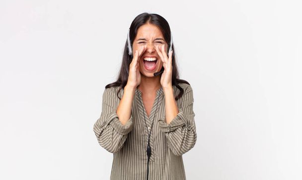 молодая испаноязычная женщина чувствует себя счастливой, раздавая громкий крик с руками рядом с ртом с наушниками. концепция телемаркетинга - Фото, изображение