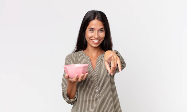 young hispanic woman pointing at camera choosing you and holding an empty bowl or pot - Foto, afbeelding