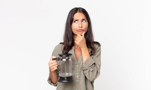 young hispanic woman thinking, feeling doubtful and confused and holding a manual coffee maker - Foto, Imagen