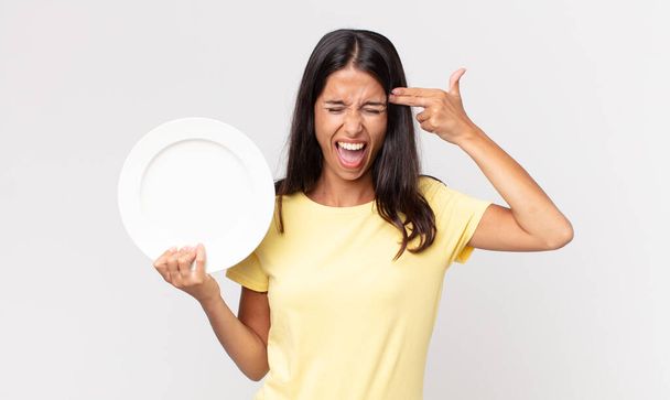 young hispanic woman looking unhappy and stressed, suicide gesture making gun sign and holding an empty plate - Foto, immagini