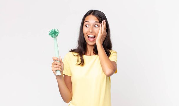 young hispanic woman feeling happy, excited and surprised and holding a dish cleaning brush - Fotografie, Obrázek