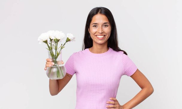 young hispanic woman smiling happily with a hand on hip and confident holding decorative flowers - Photo, Image