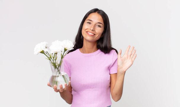 nuori latino nainen hymyilee iloisesti, vilkuttaa käsi, toivottaa tervetulleeksi ja tervehtii sinua tilalla koriste kukkia - Valokuva, kuva