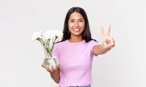 young hispanic woman smiling and looking friendly, showing number two holding decorative flowers - Fotó, kép