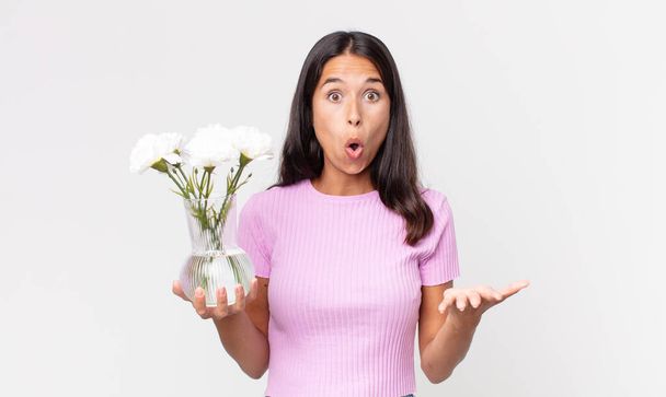young hispanic woman amazed, shocked and astonished with an unbelievable surprise holding decorative flowers - Photo, Image