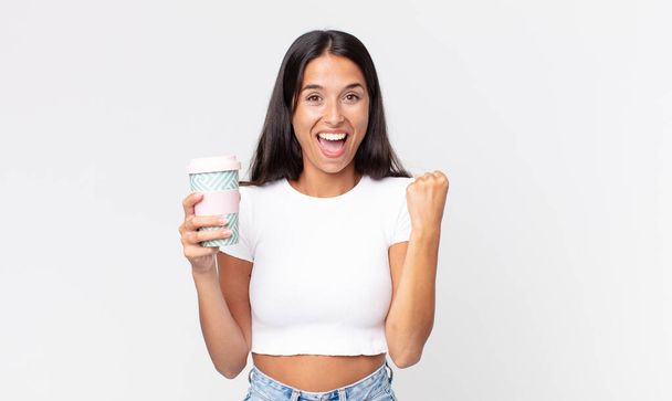 young hispanic woman feeling shocked,laughing and celebrating success and holding a take away coffee container - Foto, imagen