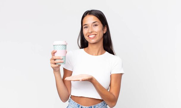 young hispanic woman smiling cheerfully, feeling happy and showing a concept and holding a take away coffee container - Photo, Image