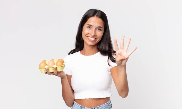 young hispanic woman smiling and looking friendly, showing number four and holding an eggs box - Foto, Imagen