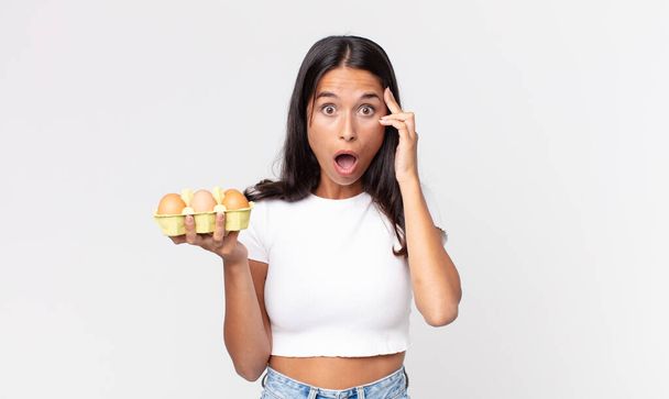 young hispanic woman looking surprised, realizing a new thought, idea or concept and holding an eggs box - Фото, изображение