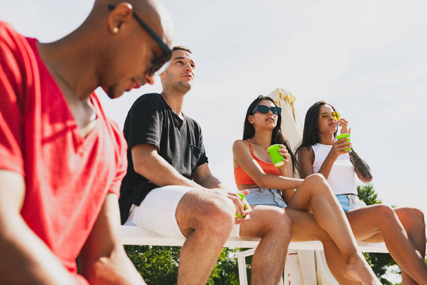 Bir grup genç, erkek ve kadın plajda dinleniyor, soğuk içecekler içiyor, eğleniyor, güneşleniyor. - Fotoğraf, Görsel