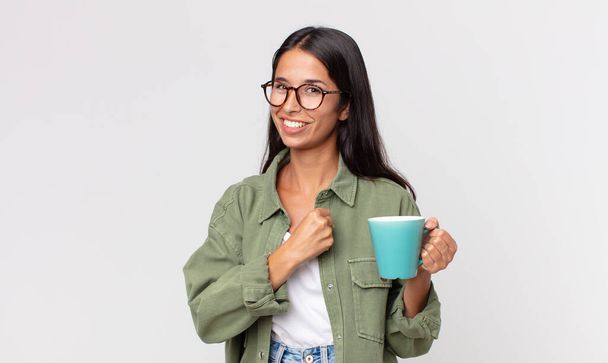 young hispanic woman feeling happy and facing a challenge or celebrating and holding a coffee mug - Foto, Bild