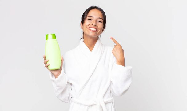 young hispanic woman smiling confidently pointing to own broad smile wearing bathrobe and holding a shampoo - Photo, image