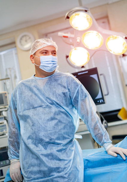 Surgeon in scrubs and face mask standing in operating theatre and looking away. Portrait of male doctor in modern medical room - Foto, Bild