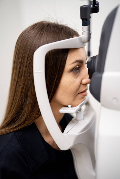 Brunette woman has eyes examined on a slit lamp. Modern equipment for eye sight check up in clinic. Closeup - 写真・画像