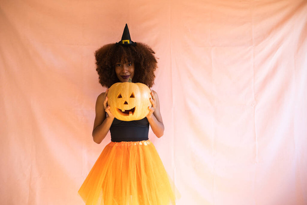 giovane donna afroamericana vestita da strega per la festa di Halloween e tenendo in mano una zucca. Indossa un cappello da strega e una gonna arancione. La donna sorride alla telecamera con la zucca. - Foto, immagini