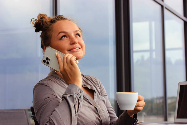 Eine verträumte Geschäftsfrau vor modernen Panoramafenstern in einem Café, auf der Restaurantterrasse und im Gespräch mit dem Smartphone. Nettes Mädchen im grauen Businessanzug, das vom Laptop aus ferngesteuert arbeitet, plaudert, Kaffee trinkt. - Foto, Bild