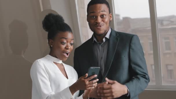 Young excited amazed African colleagues looking at smartphone screen, feel overjoyed euphoric with mobile online bet bid game app win. Businesswoman and businessman celebrate good news victory concept - Footage, Video