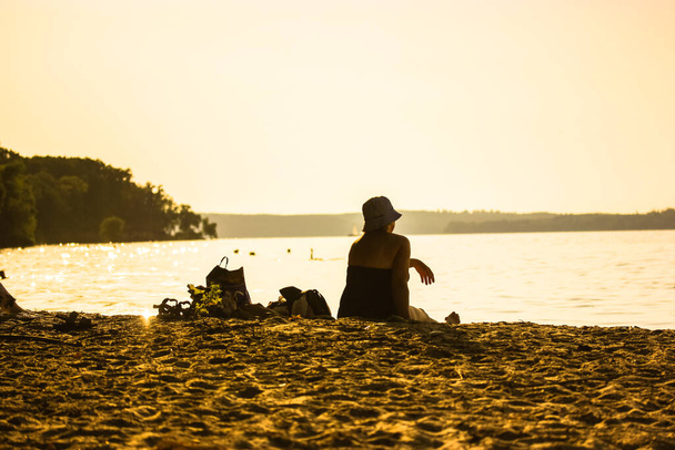 パナマの女性は、日没時に川、湖、海の砂州の銀行に座ってリラックス。バックビュー。リラックスした女性のシルエットを屋外で。美しい穏やかな風景です。黄色のトーン。夏休み旅行のコンセプト. - 写真・画像