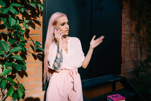Caucasian tattooed hipster girl with colored pink hair puzzled on received information during international cellphone call, unhappy female connecting to 4g in roaming for phoning to customer service - Photo, image