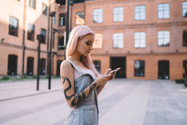 Side view of tattooed hipster teenager typing sms message for sending via mobile application, millennial youngster with pink hair using 4g wireless internet for browsing web page and install app - 写真・画像
