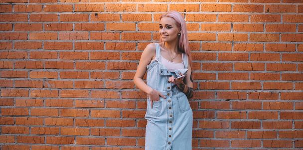 Carefree female millennial holding modern cellphone device standing at urbanity with promotional brick wall on background, happy hipster girl dressed in jeans sundress smiling at publicity area - Foto, afbeelding