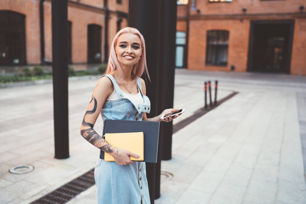 Cheerful tattooed artist with sketchbook and notepad looking away and smiling at urban setting, happy Caucasian hipster girl with tattooed hand holding modern smartphone technology for blogging - Photo, Image