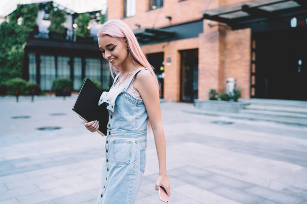 Happy pink haired female designer 20 years old holding education sketchbook walking at urbanity and smiling, cheerful Caucasian woman dressed in trendy denim apparel enjoying time at city street - Фото, зображення